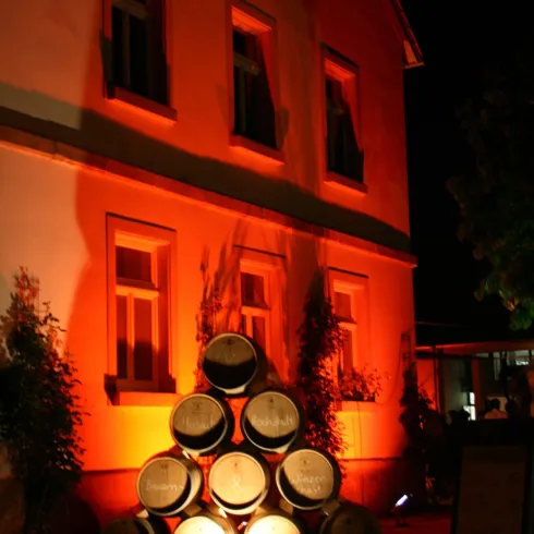 Ein Gebäude bei Nacht, beleuchtet in einem warmen Orangeton. Im Vordergrund steht ein Turm aus Holzfässern.