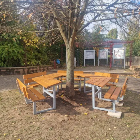 Ein angenehmer Sitzbereich aus Holz mit einem Tisch und Bänken unter einem Baum. Die Umgebung ist grün und einladend, ideal für gesellige Treffen.