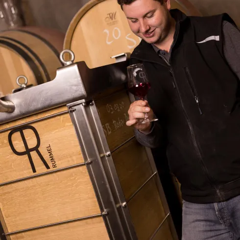 Ein Mann steht neben einem großen Weinfass und hält ein Glas Wein in der Hand. Er schaut lächelnd auf den Wein und befindet sich offensichtlich in einem Weinkeller.