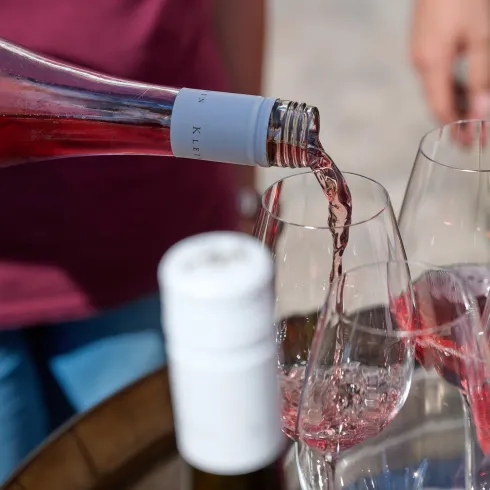 Eine Flasche Roséwein wird in Gläser gegossen. Im Hintergrund sind Menschen zu sehen, die das Getränk genießen.