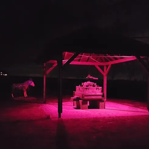 Eine beleuchtete Holzlaube mit einer roten Lichtquelle steht in der Dunkelheit. Im Hintergrund ist ein Pferd aus Holz zu sehen.