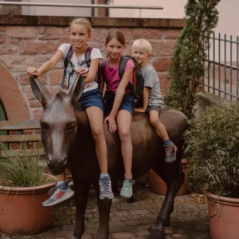 Drei Kinder sitzen auf einer Eselfigur in einem Innenhof. Im Hintergrund sind Pflanzen und eine Mauer zu sehen.
