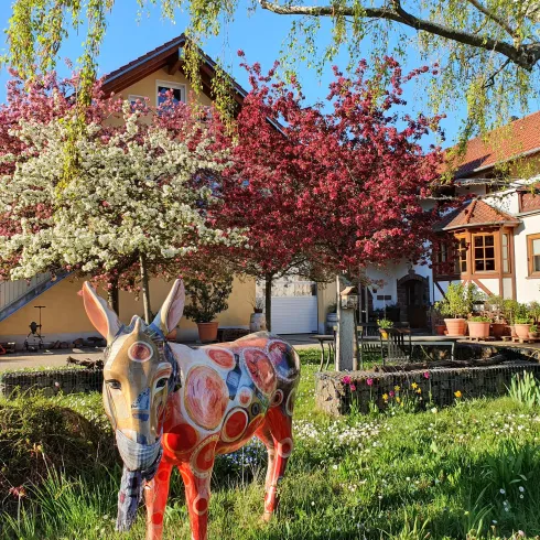 Ein buntes Esel-Bild steht auf einer Wiese vor einem schönen Haus. Im Hintergrund blühen pinke und weiße Bäume.