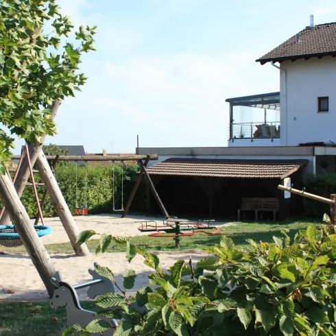 Ein Spielplatz mit Schaukeln und einer Rutsche hinter einem Haus. Die Umgebung ist grün und einladend.