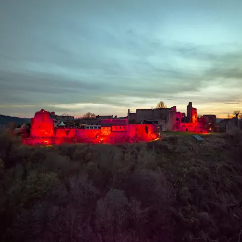 Eine Burg, die bei Sonnenuntergang in rot beleuchtet ist, steht auf einem Hügel. Umgeben von Bäumen und einem bewölkten Himmel strahlt sie eine mystische Atmosphäre aus.