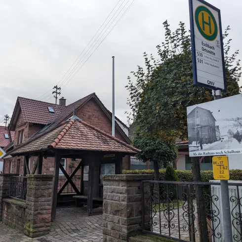 Eine Bushaltestelle mit einem überdachten Wartebereich und einem historischen Schild. Im Hintergrund sind mehrere Häuser mit roten Dächern zu sehen.