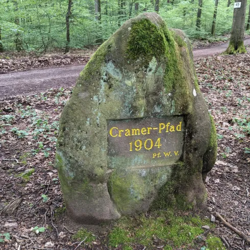 Ein großer, moosbedeckter Felsen mit einer beschrifteten Tafel steht im Wald. Die Tafel zeigt „Cramer-Pfad 1904“.