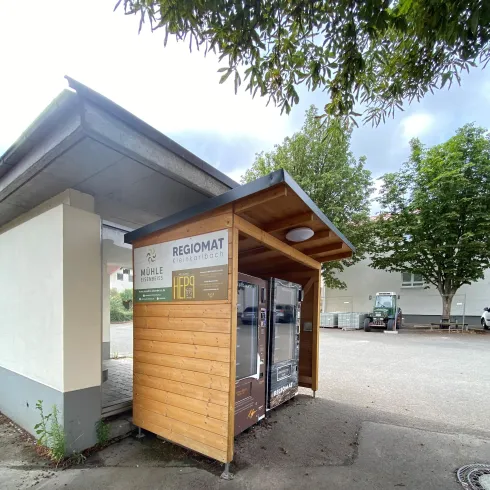 Ein Regiomat aus Holz steht an einem ruhigen Ort. Er bietet Produkte aus der Region an und ist von Bäumen umgeben.