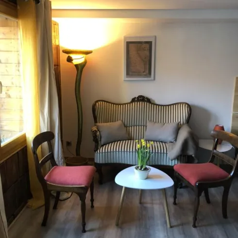 Ein gemütliches Wohnzimmer mit einem bequemen Sofa und zwei Stühlen. Ein kleiner Tisch mit einer Pflanze steht in der Mitte, und warmes Licht schafft eine einladende Atmosphäre.