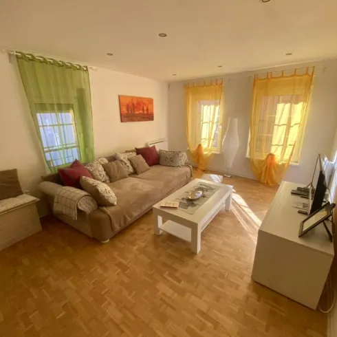 Ein helles Wohnzimmer mit einem bequemen Sofa und bunten Vorhängen. Ein Couchtisch und ein Fernseher ergänzen die Einrichtung.
