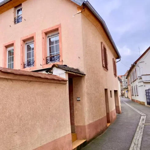 Ein kleines, charmantes Haus in einer schmalen Gasse. Die Fassade ist in sanften Rosatönen gehalten und hat große Fenster.