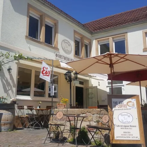 Ein einladendes Café mit Außenbereich und Sonnenschirmen. Die Fassade ist hell und freundlich, ideal für einen entspannten Aufenthalt.