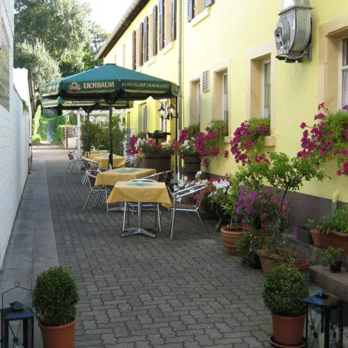 Eine charmante Gasse mit gelben Wänden und blühenden Pflanzen. Tische und Stühle unter einem Sonnenschirm laden zum Verweilen ein.