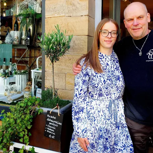 Ein Mann und eine Frau stehen lächelnd vor einem Ladengeschäft. Im Hintergrund sind Pflanzen und Möbel sichtbar, die zur Dekoration des Geschäfts beitragen.