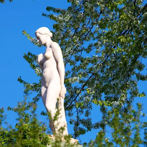 Eine elegante Statue einer nackten Frau steht vor einem strahlend blauen Himmel. Umgeben von grünen Blättern vermittelt sie eine ruhige und harmonische Atmosphäre.