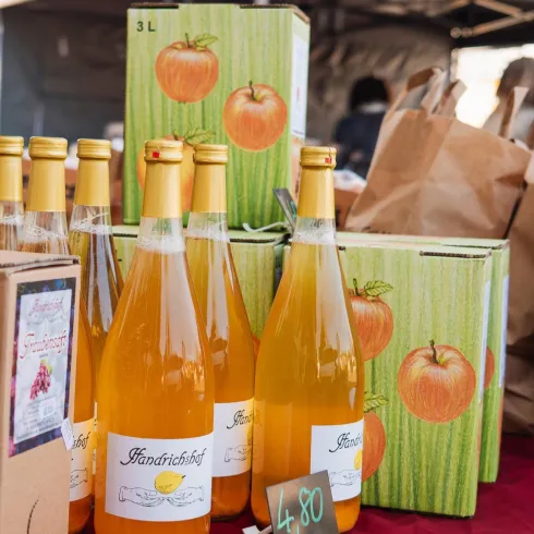 Eine Auswahl an Flaschen mit Apfelsaft und einigen Pappkartons auf einem Markt. Die Flaschen sind leuchtend orange und die Kartons zeigen frische Äpfel.