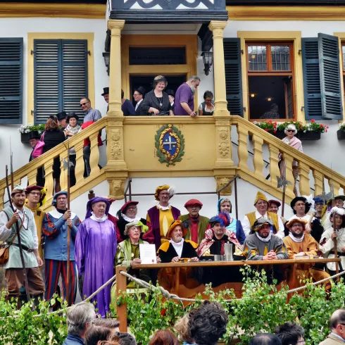 Eine Gruppe von Menschen in historischen Kostümen steht auf einer Treppe vor einem Gebäude. Einige Personen halten Holzstäbe und es gibt eine festliche Atmosphäre.