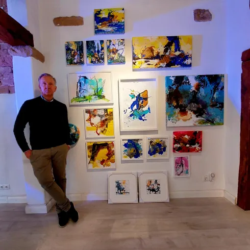 Ein Mann steht neben einer Wand mit mehreren farbenfrohen Gemälden. Die Kunstwerke zeigen verschiedene abstrakte Designs und sind in unterschiedlichen Rahmen präsentiert.