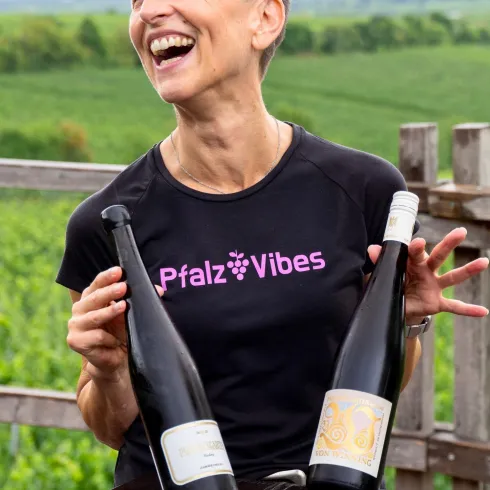 Eine fröhliche Frau hält zwei Weinflaschen in den Händen und lächelt. Im Hintergrund sind grüne Weinberge zu sehen.