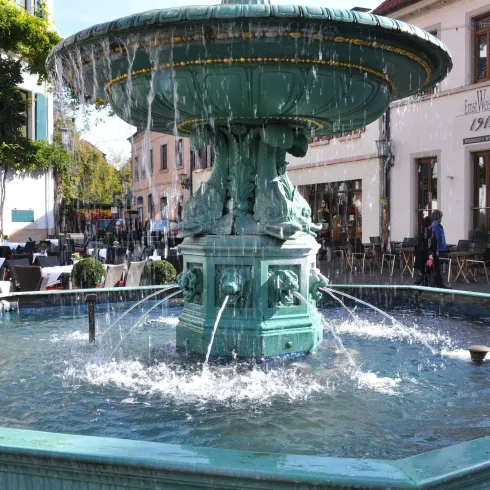 Ein schöner Brunnen mit sprudelndem Wasser in einem gemütlichen Platz. Im Hintergrund sind Cafés und Bäume zu sehen.