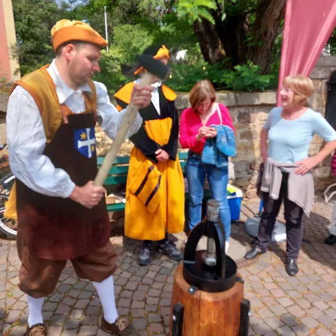 Ein Mann in historischer Kleidung zeigt eine traditionelle Handwerkskunst. Eine Gruppe von Zuschauern beobachtet interessiert.
