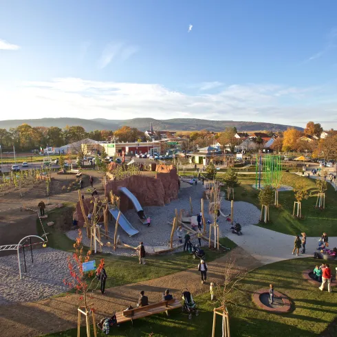 Ein großer Spielplatz mit verschiedenen Spielgeräten und vielen Kinder und Erwachsenen. Die Landschaft ist grün mit einem klaren Himmel im Hintergrund.
