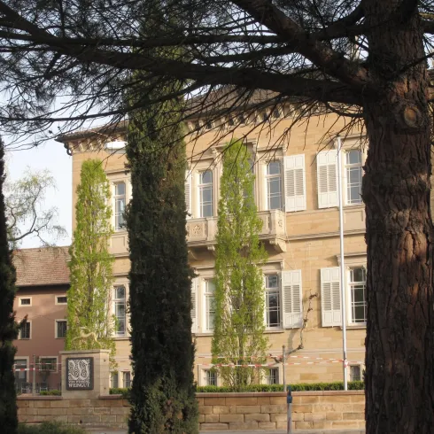 Ein historisches Gebäude umgeben von hohen Bäumen. Die Fassade ist mit Efeu bewachsen und hat viele Fenster.