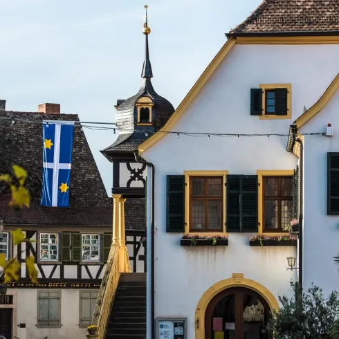 Ein historisches Gebäude mit gelben Fensterrahmen und einem hübschen Dach ist zu sehen. Im Hintergrund hängt eine Flagge mit blauem und gelbem Design.