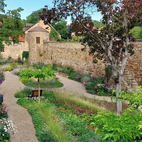 Ein schöner Garten mit bunten Blumen und grünen Pflanzen. Im Hintergrund ist eine alte Steinmauer und ein kleines Gebäude zu sehen.