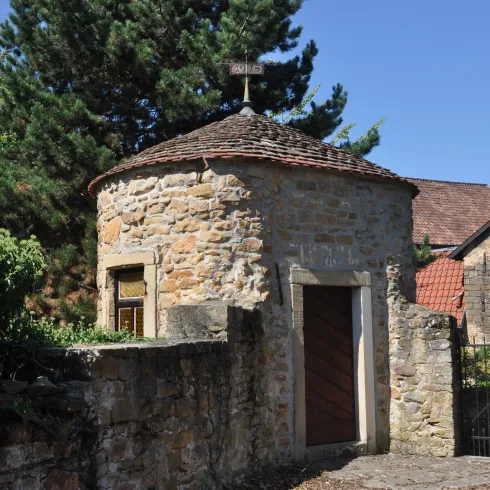 Ein historischer runder Steinbau mit einem abgedachten Dach. Umgeben von Pflanzen und einem Tor.