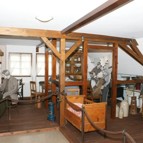 Ein traditionell eingerichteter Raum mit Holzdekor und einem Tisch. Eine Figur sitzt am Tisch, umgeben von alten Haushaltsgegenständen.