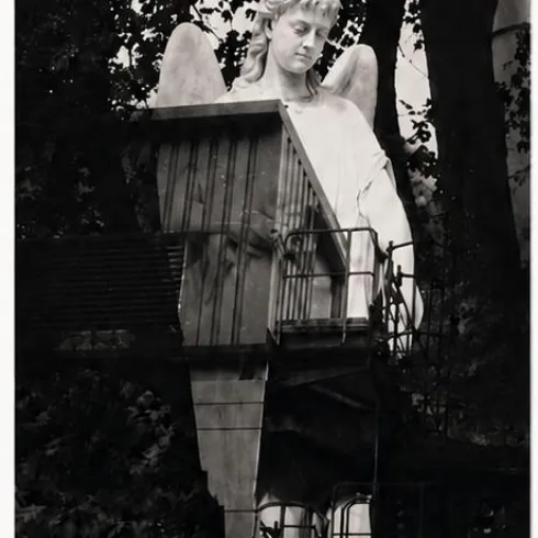Eine große Statue eines Engels mit Flügeln. Der Engel steht vor einer Struktur mit Gitter und ist von Bäumen umgeben.