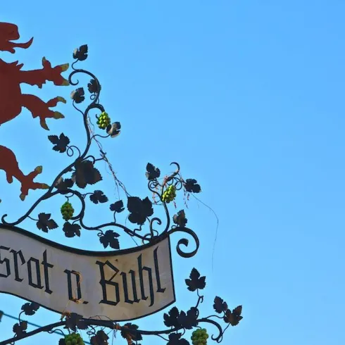 Ein kunstvoller Metallschild mit einem Löwen und Traubenmotiven. Darauf steht "Reichsrat d. Buhl" vor einem klaren blauen Himmel.