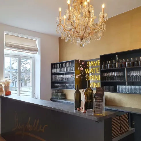Eine elegante Bar mit einem schönen Kronleuchter und einem gut gefüllten Regal für Gläser. An der Wand steht der Slogan "Save water, drink wine".