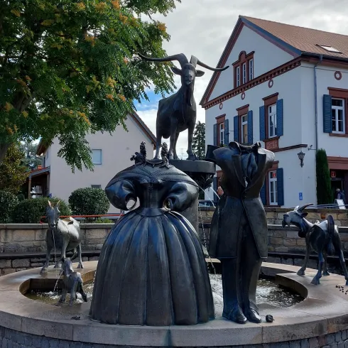 Eine kreative Brunnenskulptur mit Figuren von Menschen und Tieren, darunter eine Ziege und verschiedene andere Tiere. Der Brunnen steht vor einem historischen Gebäude in einer ruhigen Umgebung.
