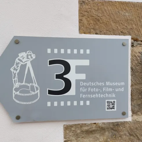 Ein Schild zeigt den Weg zum Deutschen Museum für Foto-, Film- und Fernsehtechnik. Es befindet sich an einer Wand und zeigt eine Pfeilrichtung sowie eine grafische Darstellung.