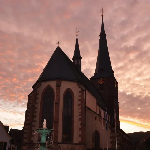 Eine alte Kirche mit zwei hohen Türmen steht vor einem bunten Sonnenuntergang. Die Wolken sind strahlend und verleihen der Szene eine malerische Atmosphäre.