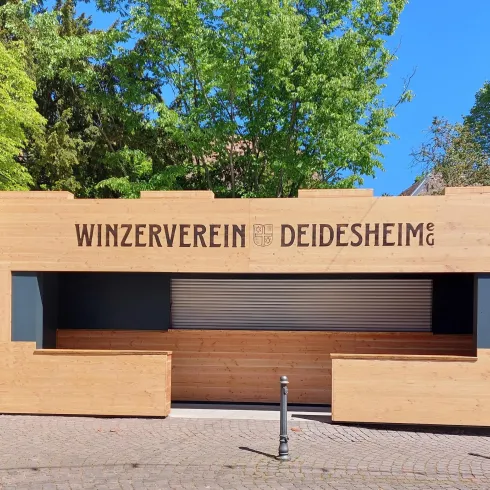 Ein modernes Gebäude mit dem Schriftzug "Winzerverein Deidesheim". Es befindet sich in einer grünen Umgebung mit Bäumen.