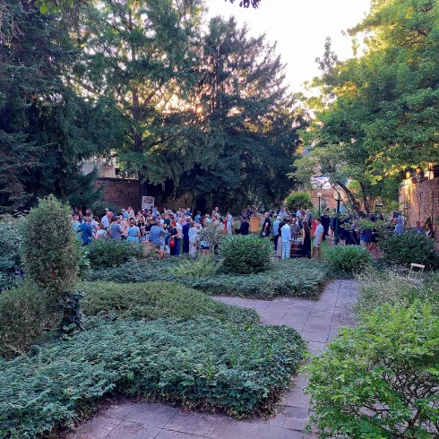 Eine große Gruppe von Menschen versammelt sich in einem grünen Garten. Bäume und Sträucher umgeben den Bereich, während der Abendhimmel erhellt wird.