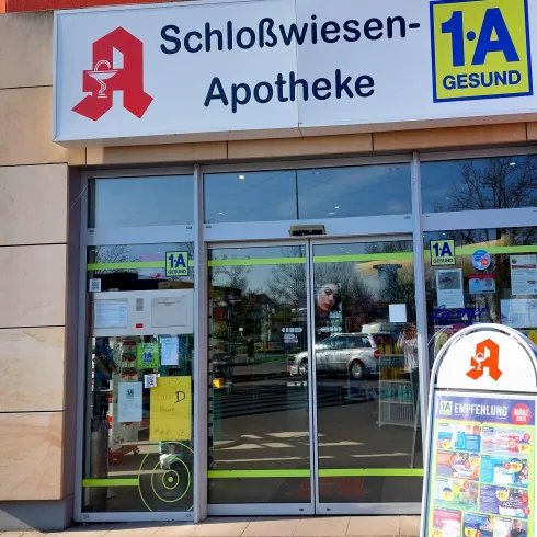Eine Apotheke mit dem Namen "Schloßwiesen-Apotheke" ist zu sehen. Der Eingang ist gut sichtbar und es gibt ein Werbeschild im Vordergrund.