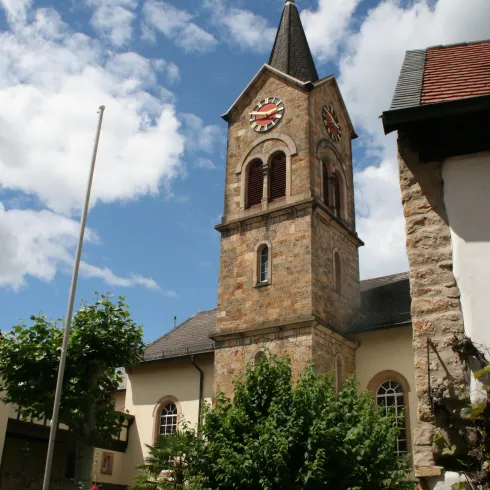 Eine alte Kirche mit einem markanten Turm und einer Uhr. Im Vordergrund sind grüne Bäume und traditionelle Gebäude zu sehen.