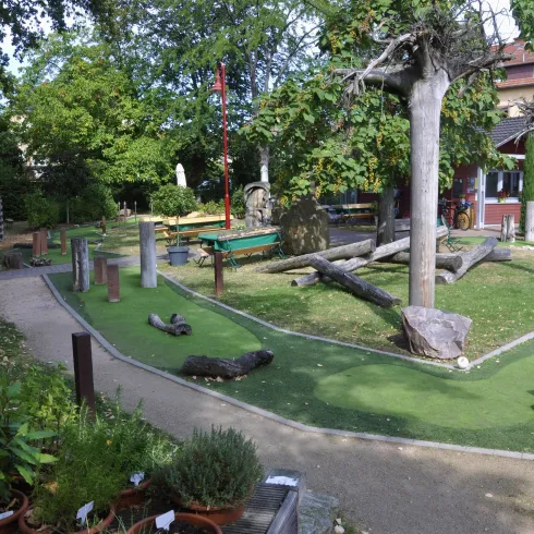 Ein schöner Minigolfplatz mit verschiedenen Hindernissen und viel Grün. Im Hintergrund sind Bäume und ein Gebäude zu sehen.