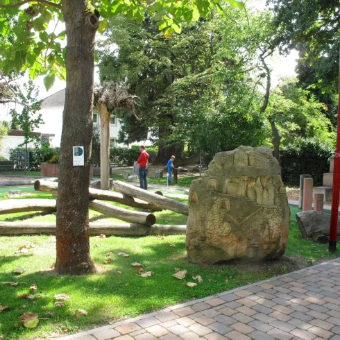 Ein schöner Park mit grünen Wiesen und Bäumen. Im Hintergrund sind Personen zu sehen, die sich auf dem Spielplatz aufhalten.
