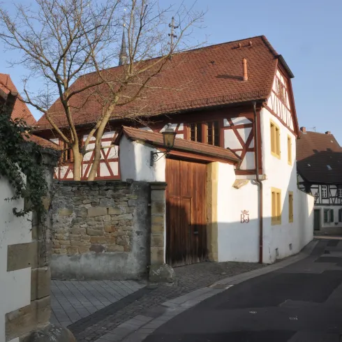 Eine ruhige Gasse mit traditionellen Fachwerkhäusern und einer alten Mauer. Bäume wachsen in der Nähe und die Straße ist gepflastert.