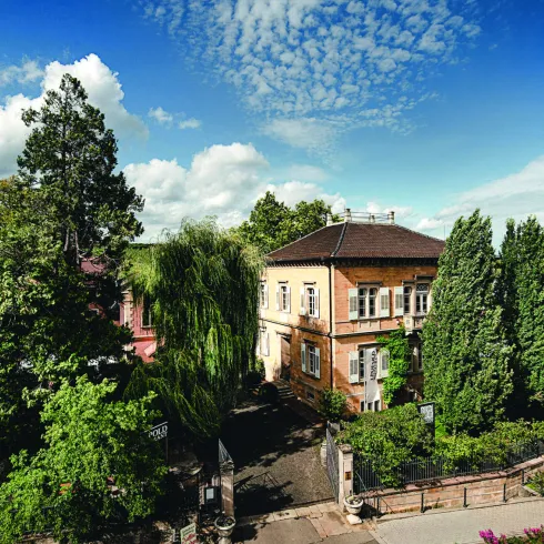 Ein historisches Gebäude umgeben von üppigem Grün und Bäumen. Der Himmel ist blau mit einigen Wolken.