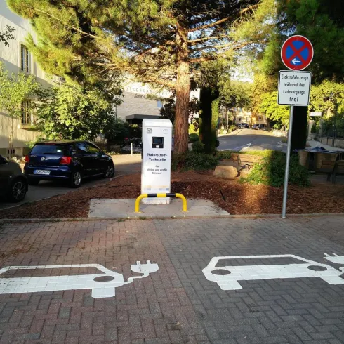 Eine Elektroladestation für Autos ist auf einem Parkplatz zu sehen. Neben der Station sind die Umrisse von zwei Elektroautos auf dem Boden markiert.