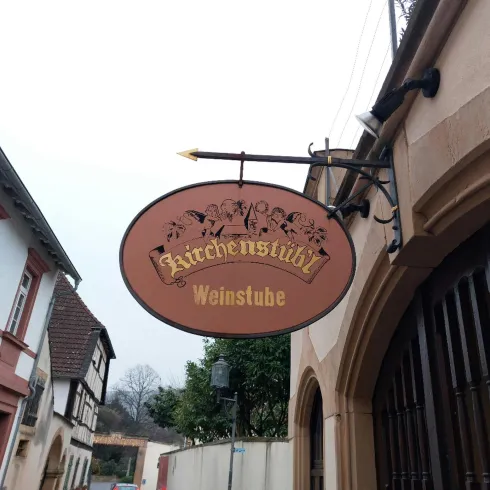 Ein gemütliches Weinschänke-Schild hängt an einem historischen Gebäude. Die Umgebung zeigt typische deutsche Architektur und eine ruhige Atmosphäre.