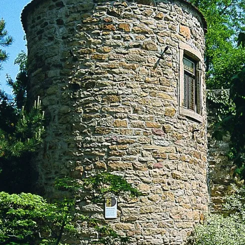 Ein runder, steinerner Turm umgeben von Bäumen und Pflanzen. Der Turm hat ein kleines Fenster und eine Statue an der Spitze.