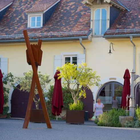 Ein charmantes Gebäude mit gelber Fassade und einem schönen Garten. Im Vordergrund steht eine große Holzskulptur und einige Sonnenschirme.
