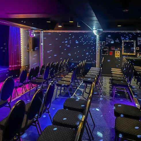 Ein kleiner Veranstaltungsraum mit Reihen von schwarzen Stühlen. Die Bühne ist beleuchtet, und es gibt ein pinkes und blaues Lichtspiel an den Wänden.